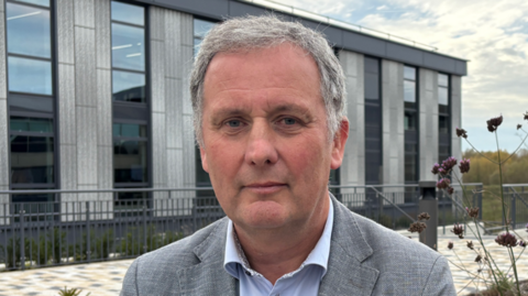 Robert Evans is in the middle looking at the camera. He has short grey hair and is wearing a blue shirt and grey suit jacket. Behind him is a grey bricked building with tall windows.