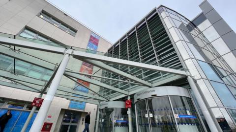 The main entrance of Broomfield Hospital in Chelmsford showing a metal and glass canopy sticking out from the front of the building which is clad with a natural coloured material with multi-coloured blocks forming a strip down the middle of the building. There are rotating doors.