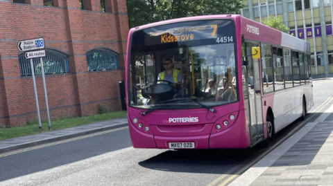 A purple and white bus with "7 Kidsgrove" on the screen at the top drives towards us along a road.