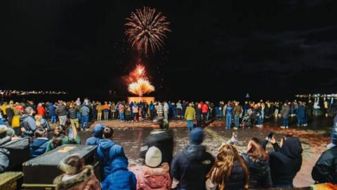 Hundreds of people in warm coats and hats stand on Douglas promenade looking out at the bay, which has red fireworks exploding.