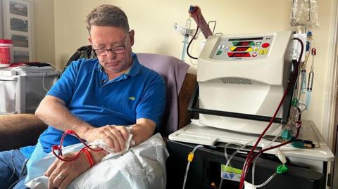 Tony Henderson sitting in his living room undergoing home dialysis. His dialysis machine is to his left and you can see tubes running into his left arm. You can see him applying pressure to his left arm with his right arm at the fistula site. Mr Henderson is wearing a royal blue polo shirt and has glasses on.