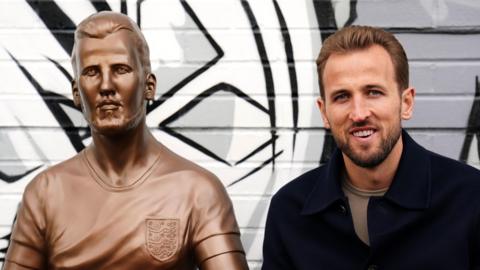 Harry Kane is seen sitting to the right of his statue. Both are seen from the chest up. The brown statue depicts him wearing an England shirt with a neutral expression while the real Harry is smiling.