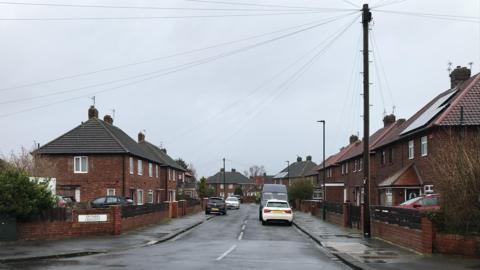 Ontario Crescent. Brick, two-storey semi-detached houses line each side of the street. They each have a slate roof. Several cars are parked along the street and a wooden telegraph pole with wires attaching to several houses stands in the foreground.