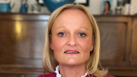 Maria Mulgrew looking to camera. She is sitting in front of a piano and is wearing a dark pink tank top and a white blouse with a frilled collar