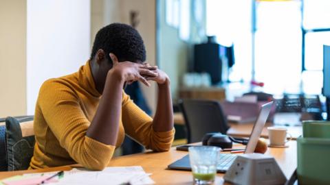 A woman in a yellow jumper holds her head in her hands as she sits at a desk 