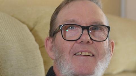 Jack Mee sits on a cream sofa and looks to the side to smile at the camera. He has short dark hair and a grey beard. He is wearing black glasses and a black jumper.