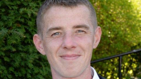 David McGuinness, man smiling and looking at camera, with green bushes in background.