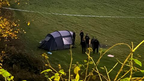 A tent and a cordon on a greenfield with trees and woodland surrounding it. There are six police officers in uniform standing nearby