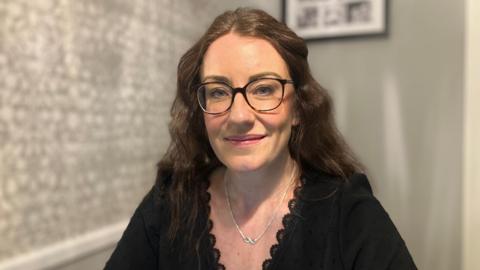 Samantha Gelder, smiles at the camera. She's wearing glasses, a black top and a silver necklace.