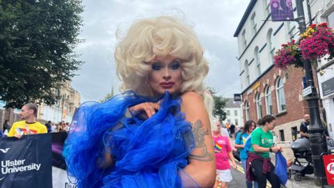 A person with blonde hair looks at the camera. They are wearing a blue piece of clothing, and they have a tattoo on their upper left arm. Behind them are a number of people.