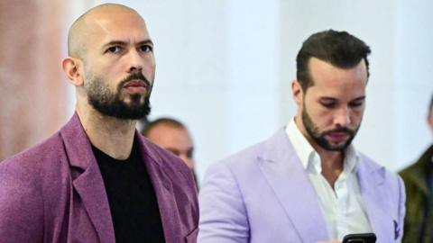 Andrew Tate on the left and Tristan in Bucharest in 2024. They both have beards. Andrew is wearing a purple jacket with black T-shirt and Tristan wears a lilac jacket and a white shirt