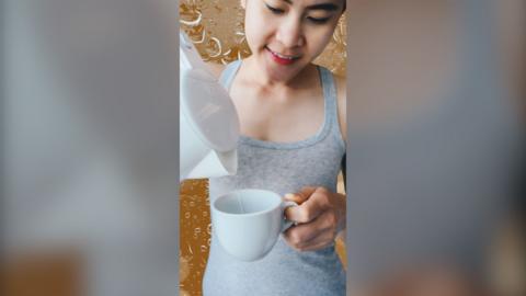 Woman wearing a grey vest top, pours water from a kettle into a mug 