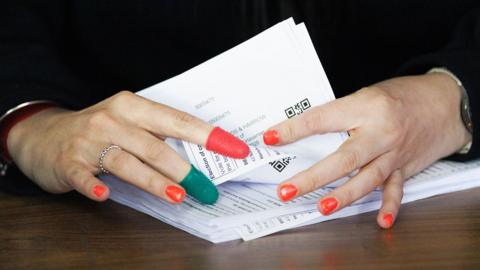 A close up image of ballot papers being counted by hands wearing thimbles on index and middle fingers and bright red nail polish, in Romford on Friday.