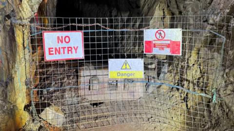 A mine shaft is blocked with a metal fence with three signs attached - one is white with red letters which say no entry. Another is white with a yellow danger symbol and the word danger. The third says no unauthorised personnel.