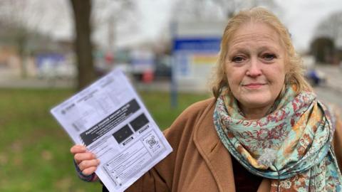 A woman with ginger hair stands outside a hospital car park holding an A4 piece of paper with details of a parking fine. She is wearing a fawn-coloured coat and a colourful scarf with floral print