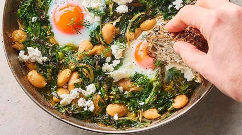 A hand dips bread into a dish of green baked eggs with butter beans