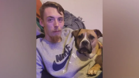 Charlie Greaves smiles at the camera while hugging a dog. He is wearing a grey sweatshirt. 