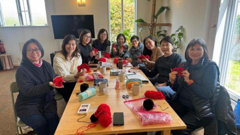 People sat around a table smiling widely holding red poppies with yarn and mugs on the table.