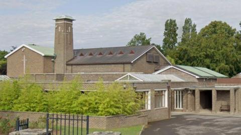 The picture show Porchester Crematorium, a building with Christian crosses imprinted onto the brickwork. Part of the building is a tall tower that rises above the rest of it. In front of the building is a lush green bush. 
