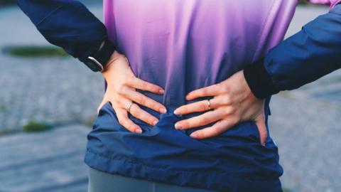 A woman holding her lower back, viewed from behind, wearing navy and purple jumper.