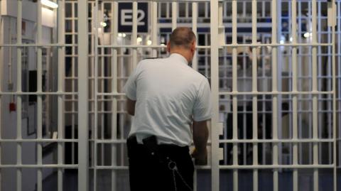 An image of a series of security gates in a prison - with floor-to-ceiling white metal bars. Above the first set it says "E/F Wing" and a prison officer - a bald man in a white shirt and black trousers - is opening the gate. We see him from behind. 