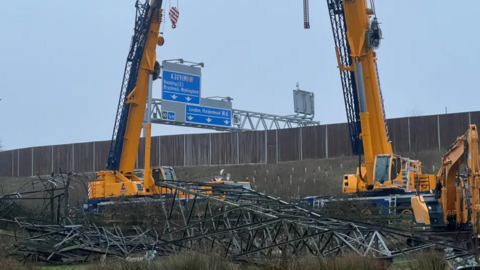 A general view of two large cranes over a pylon that has been brought down. The M4 sign is visible behind it on a grey day.