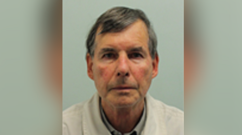 A mugshot of a man with brown hair, a white jacket and grey shirt.