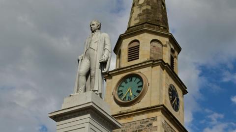 The statue and plinth of Sir Walter Scott is grey in colour. The figure of Sir Walter Scott is wearing a waistcoat, breeches and a gown. The statue stands outside the courthouse in Selkirk. The stone-built courthouse has a clock tower.