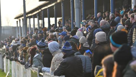 Barrow Raiders rugby fans watching their team in the stands of a stadium. They can be seen from behind and are wearing warm clothes, hats and scarves.