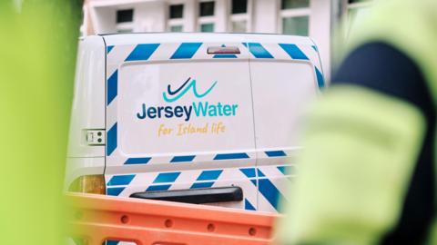 A Jersey Water white van with the company's logo on the back door is parked on a street. It is next to some orange fencing. Two people in high-vis jackets are in the foreground. Both are blurred.