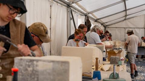 A group of people stand along a table carving stones with hammers under a marquee. There is a lot of dust everywhere.