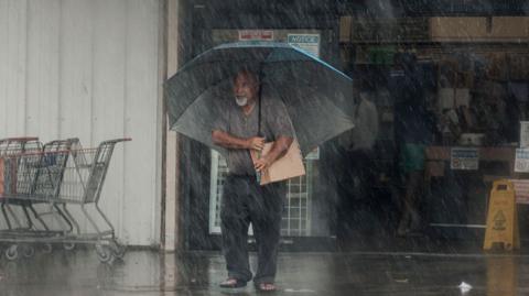 An elderly man clutching a box under his arm as he holds an umbrella over his head in very heavy rain outside of a supermarket