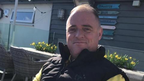 Raymond McDonald, who is balding and wearing a black gilet and yellow t-shirt, is sitting outside with daffodils in planters behind him, and the wooden wall of a building behind that. On the wall are wooden finger post signs with place names on them.