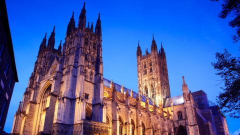 A grand cathedral illuminated with yellow lighting against a pale blue sky, with the main entrance on the left side. There are leafy branches peaking from the right-hand-side of the frame.