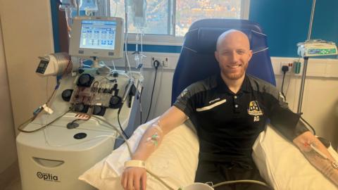 Alex Overton sits in a chair in hospital connected to a series of drips, with medical equipment to the side of him. He is smiling. 