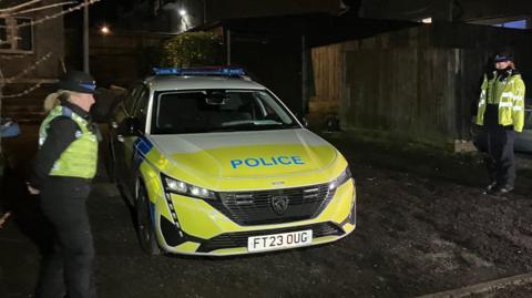 Two police officers stand either side of a police car. It is dark and they are dressed in winter uniform.