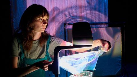 Katie-anna Whiting drapes a plastic sheet on part of an overhead projector to create an image on a wall behind her. She has long brown hair and wears a T-shirt with dungarees. 