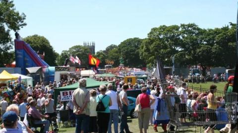 Crowds of people enjoy festival activities on a grassy field, including a fun fair