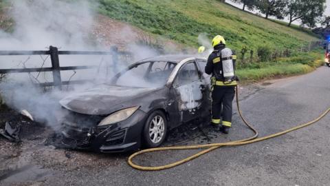 Two firefighters tackle the flames from a black car which is on fire on the side of a road next to a grass verge. Smoke is billowing out of the vehicle and scorch marks are on its door and roof.