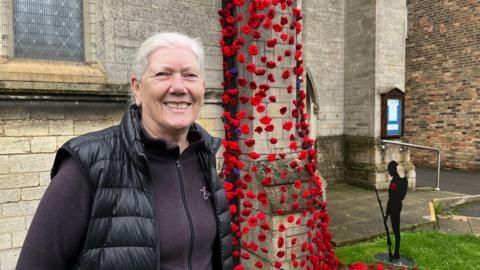 Maureen McKenna is smiling and looking at the camera to the left of the image. She is wearing a dark fleece with a zip and a black gilet over the top. Behind her is an outside wall of St Matthew's Church in Eye with black netting over a buttress covered with red and purple knitted poppies. To the right of it is a free standing black metal figure of a soldier with his head bowed. 