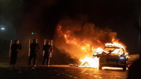 Garda officers in riot gear stand with shields deployed close to a burning garda car in Dublin on 23 November 2023