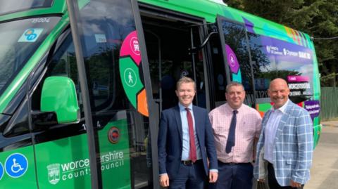 Three men standing in front of a green minibus. They are wearing ties and jackets.