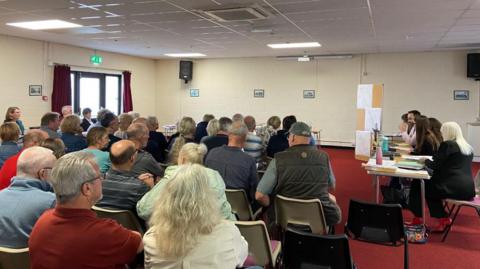 A room full of people sitting on chairs at a public inquiry, with desks and more people at the sides