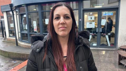 Joanne Pritchard is standing outside the Hope Centre’s day facility on a wet pavement, with the curved glass frontage of the building visible behind them. The entrance doors, noticeboards and interior lights can be seen through the windows, where people are gathered inside the warm space. The person is dressed in a winter coat suitable for the cold, rainy weather, and the surroundings include a small seating area and the brickwork of nearby buildings, reflecting the busy, urban setting of the support hub.