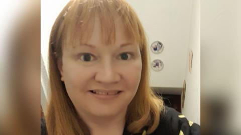 A woman with light ginger hair is taking a selfie. She appears to be standing in a staircase with pictures on a plain white wall.