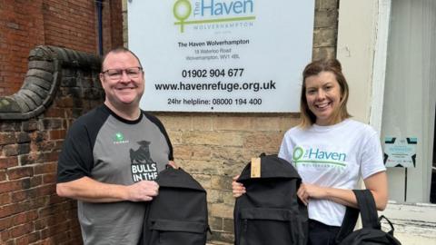 A man and a woman stand in front of the The Haven head office in Wolverhampton. Both of them are holding black backpacks and smiling at the camera.