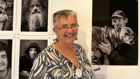Mary Mustoe smiles at the camera as she stands in front of the photos being exhibited. They are black and white and documentary style photos of men. On the right a man with an Adidas baseball cap holds a dog in his arms. On the top left, a man with a long beard looks at the camera.