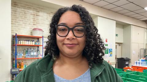 Sarah Edwards is pictured with a closed mouth smile, wearing large circular black framed glasses, a green fleece jacket and blue jumper. She has black tight curly hair. In the background are green boxes and a shelf with various drinks bottles on it.
