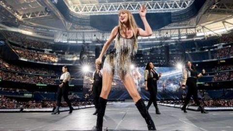 Taylor Swift stands dancing at front of stage with some backing dancers behind her surrounded by thouands in the crowds at the stadium