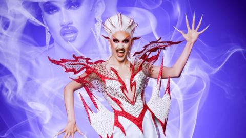 Kyran Thrax wearing a red and white outfit. The outfit has a red border which look like flames coming off the outfit. They have long nails and dramatic makeup. 